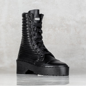 A single black lace-up ankle boot with a ribbed textured shaft, chunky platform sole, and a small silver BLAMO label on the tongue, set against a textured gray background.