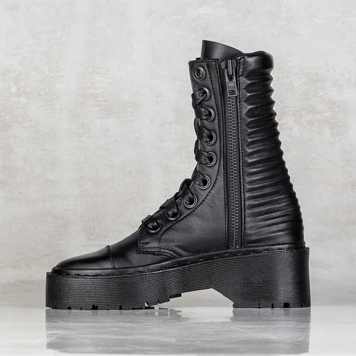 A black leather lace-up combat boot with a thick platform sole, ribbed side panels, and a side zipper, shown against a textured light gray background.