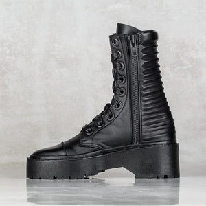 A black leather lace-up combat boot with a thick platform sole, ribbed side panels, and a side zipper, shown against a textured light gray background.