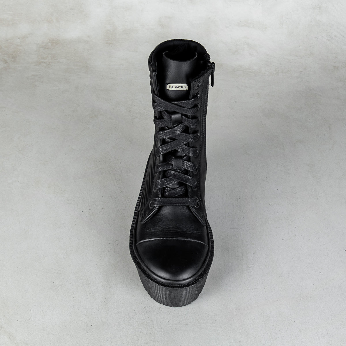 A single black leather lace-up ankle boot with a side zipper, shown from a top-down perspective on a textured light gray background.