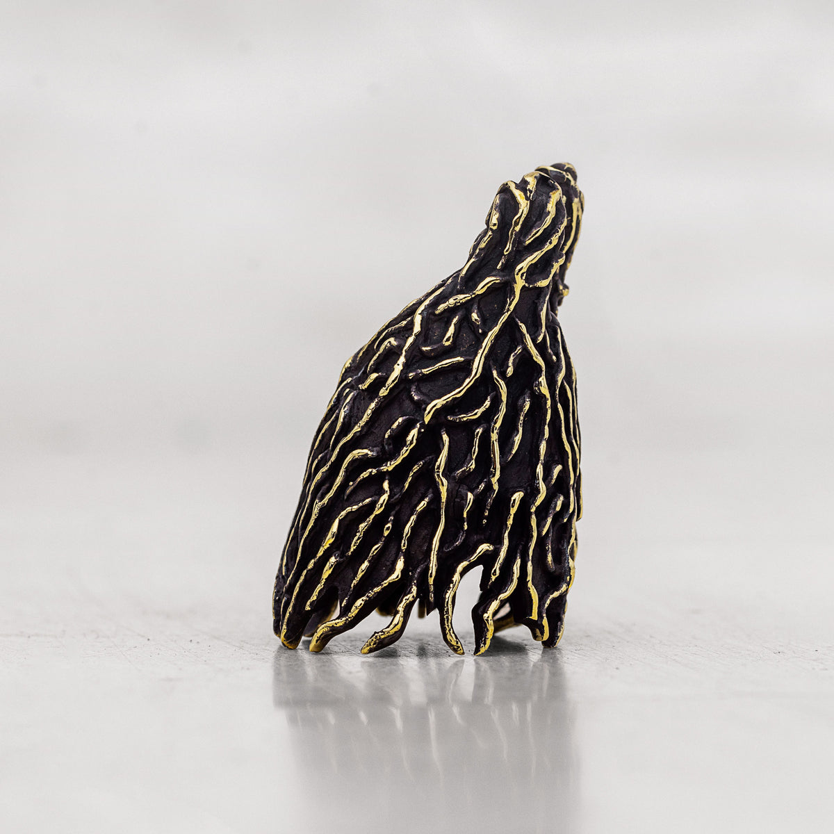 A small sculptural figurine shown from the side textured with black and gold patterns standing on a reflective light gray surface against a neutral gray background.