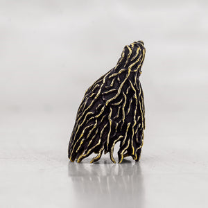 A small sculptural figurine shown from the side textured with black and gold patterns standing on a reflective light gray surface against a neutral gray background.