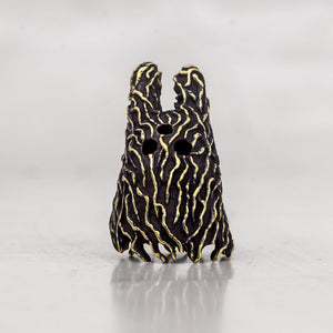 A small black and gold abstract sculpture with a textured, organic wavy pattern and three circular cutouts, photographed on a light gray reflective surface.