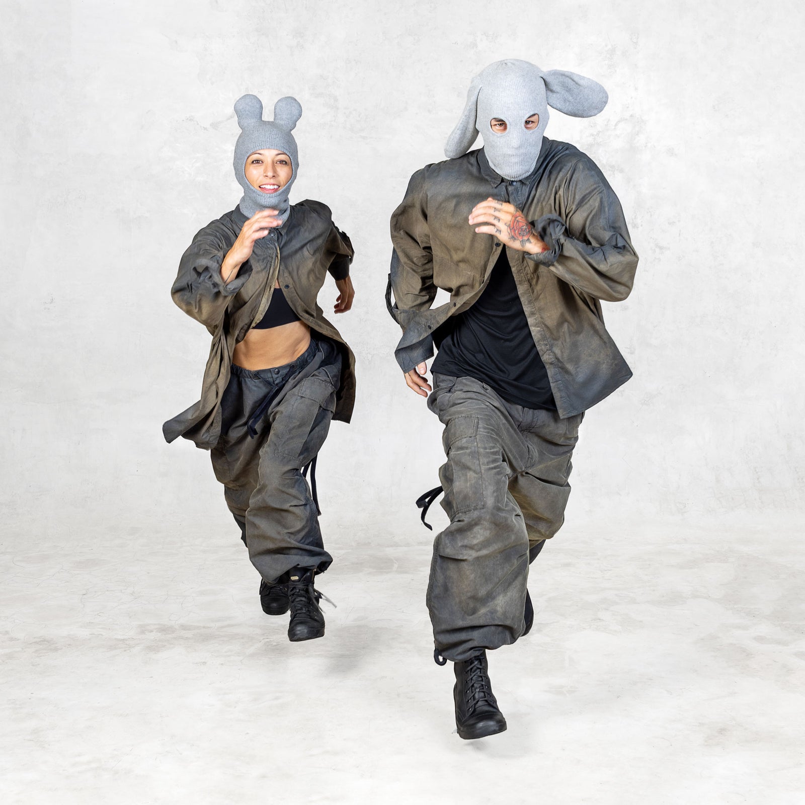 Two people wearing green and black outfits with gray animal masks in a running motion against a light gray background 