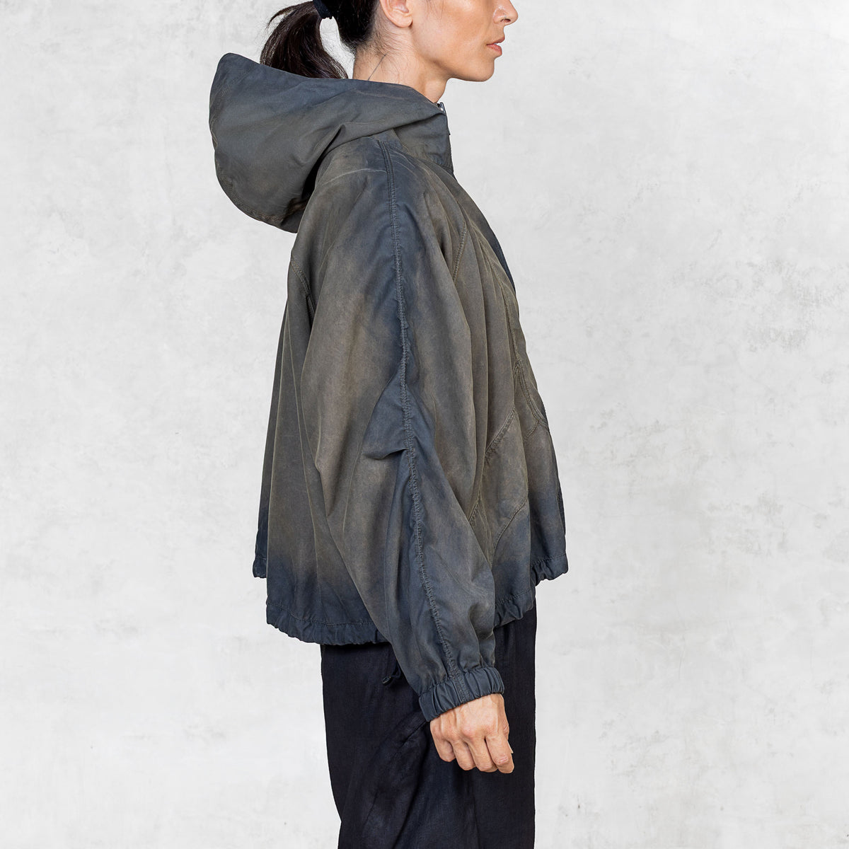 Side view of a person wearing a hand painted olive windbreaker jacket and black pants against a white background. 