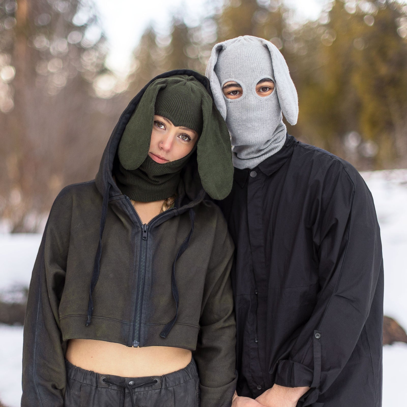 Two people wearing animal-themed knit balaclava masks and green and black outfits in a snowy forest setting