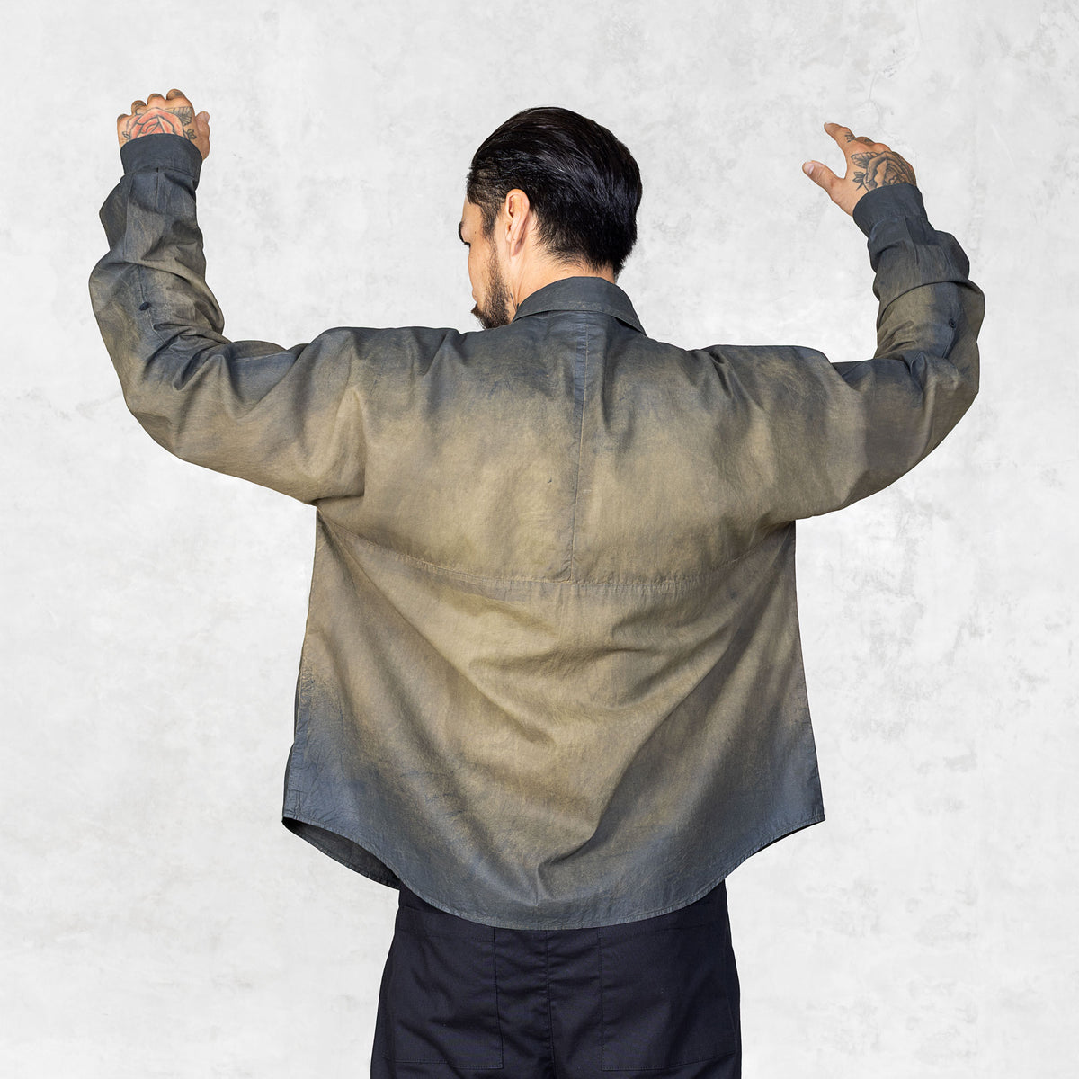 Backwards facing person with arms raised  looking off to the side wearing a hand-painted olive oversized  button up shirt and black pants against a white backdrop.