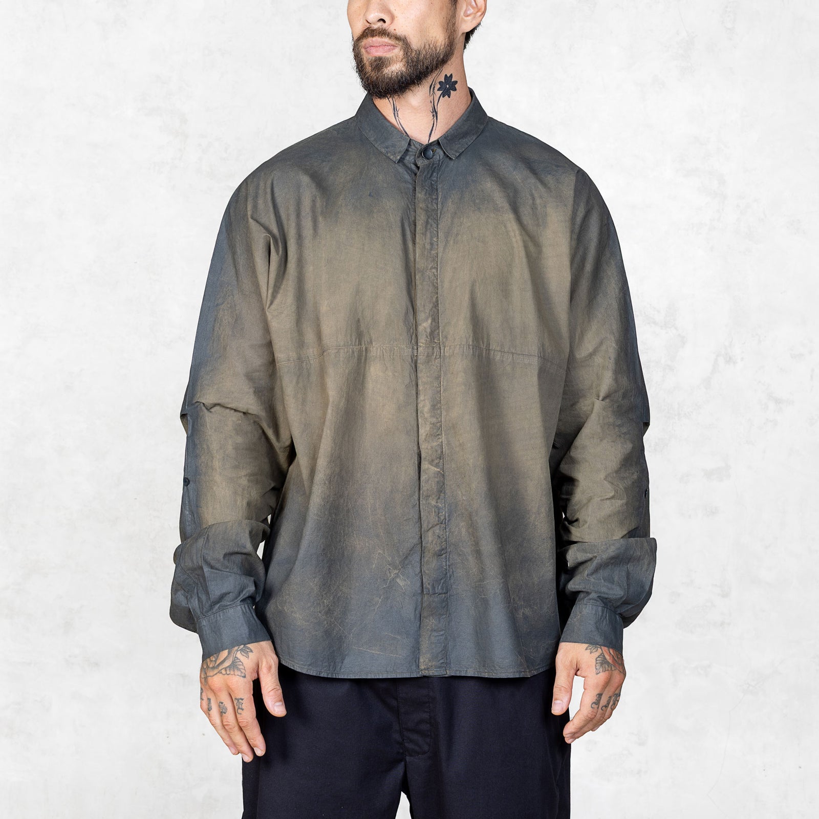 Front facing view of a bearded and tattooed person wearing a hand-painted olive oversized button up shirt and black pants against a white backdrop.