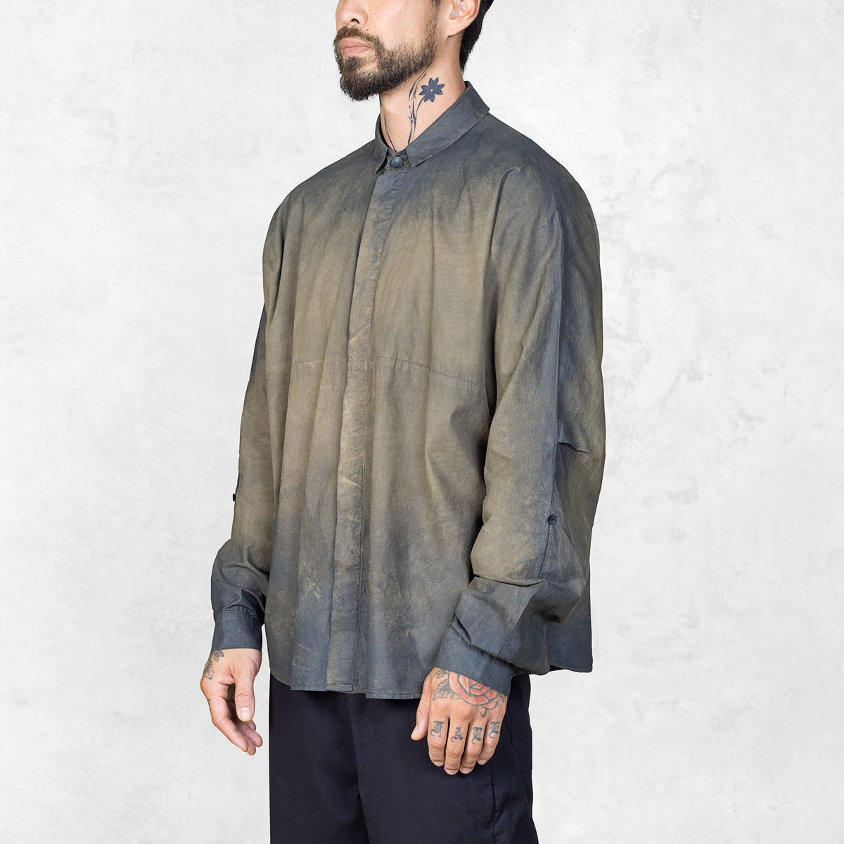 Slightly side facing view of a person wearing a hand-painted olive oversized button up shirt and black pants against a white backdrop.