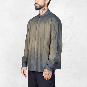 Slightly side facing view of a person wearing a hand-painted olive oversized button up shirt and black pants against a white backdrop.