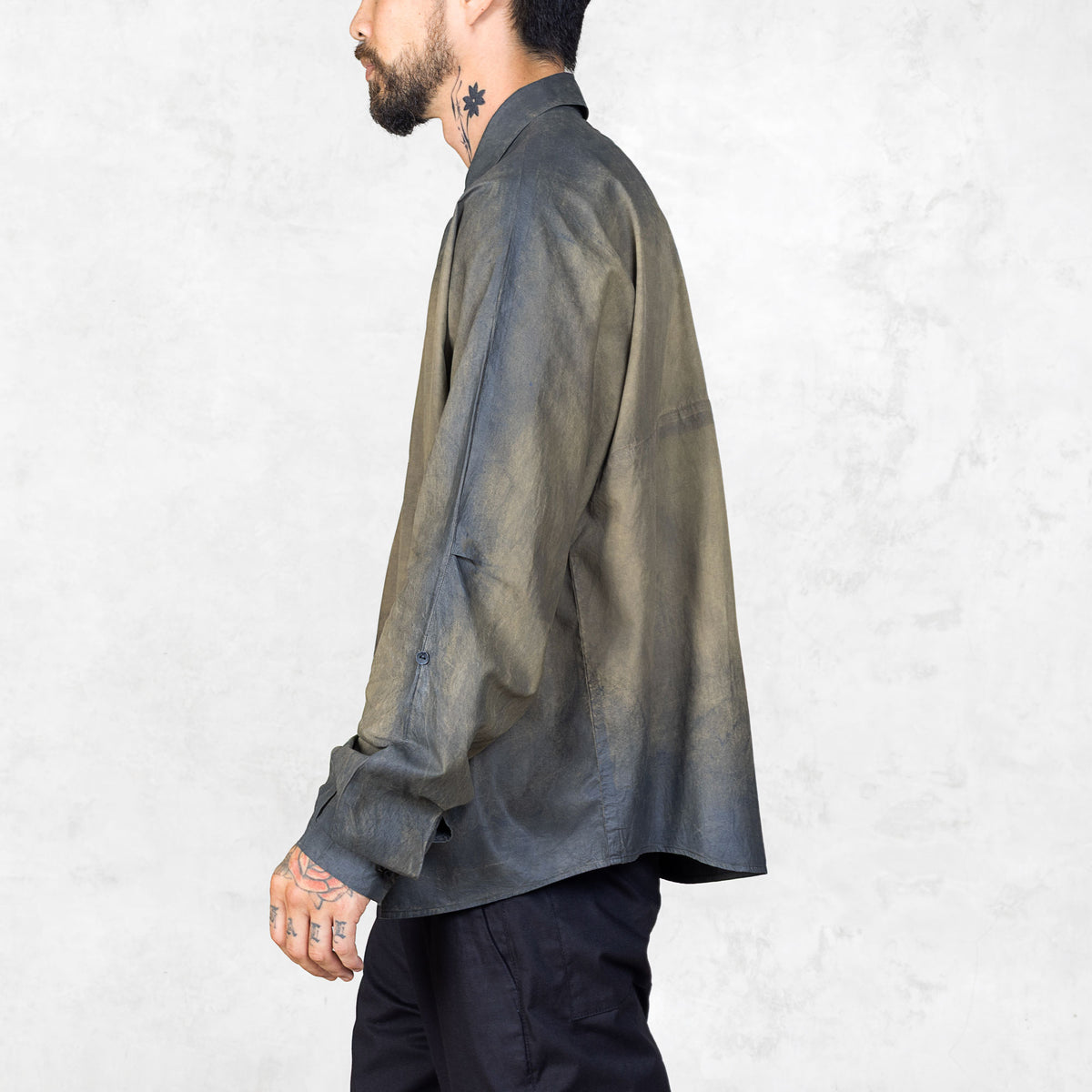 Side view of a person with neck tattoos wearing a hand-painted olive oversized  button up shirt and black pants against a white backdrop.