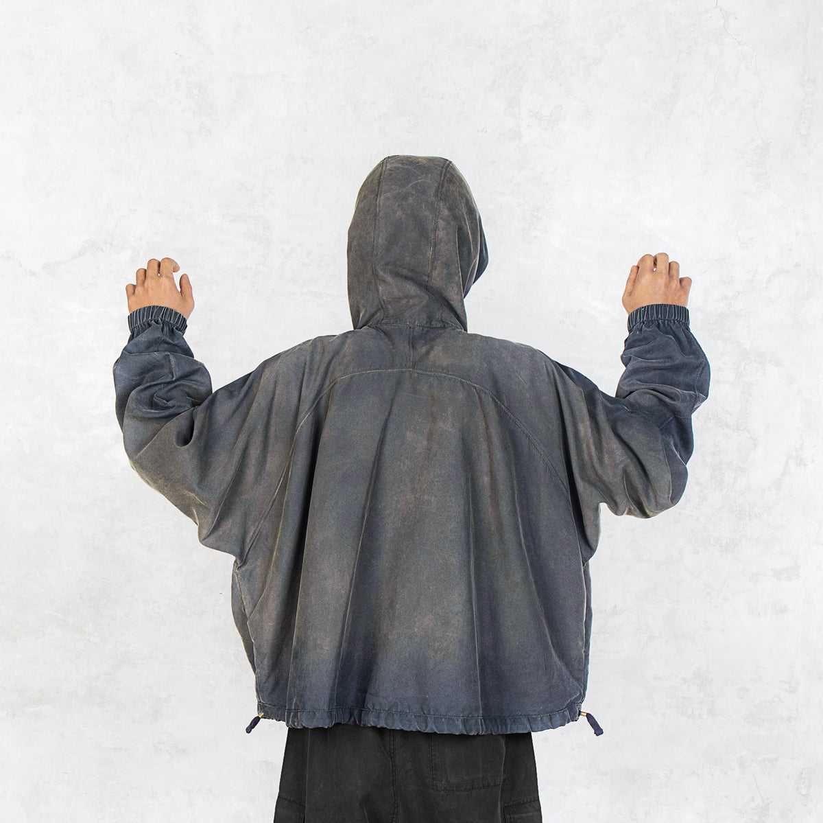 Person facing backwards with their arms raised wearing a dark olive zip up hooded jacket against a light grey background.