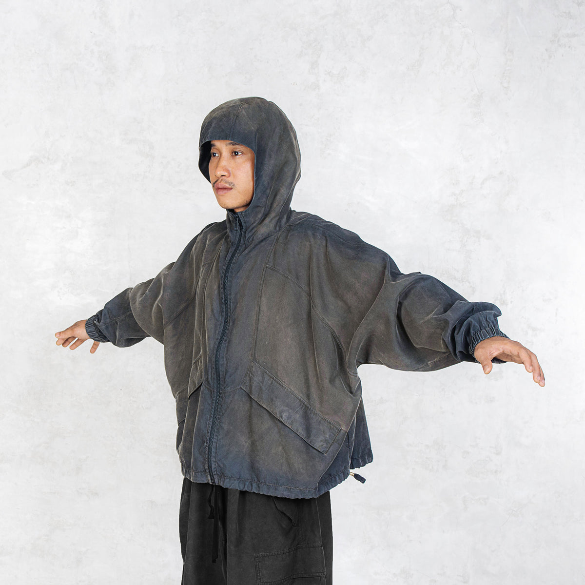 Person facing off to the side with their arms out wearing a dark olive zip up hooded jacket against a light grey background.