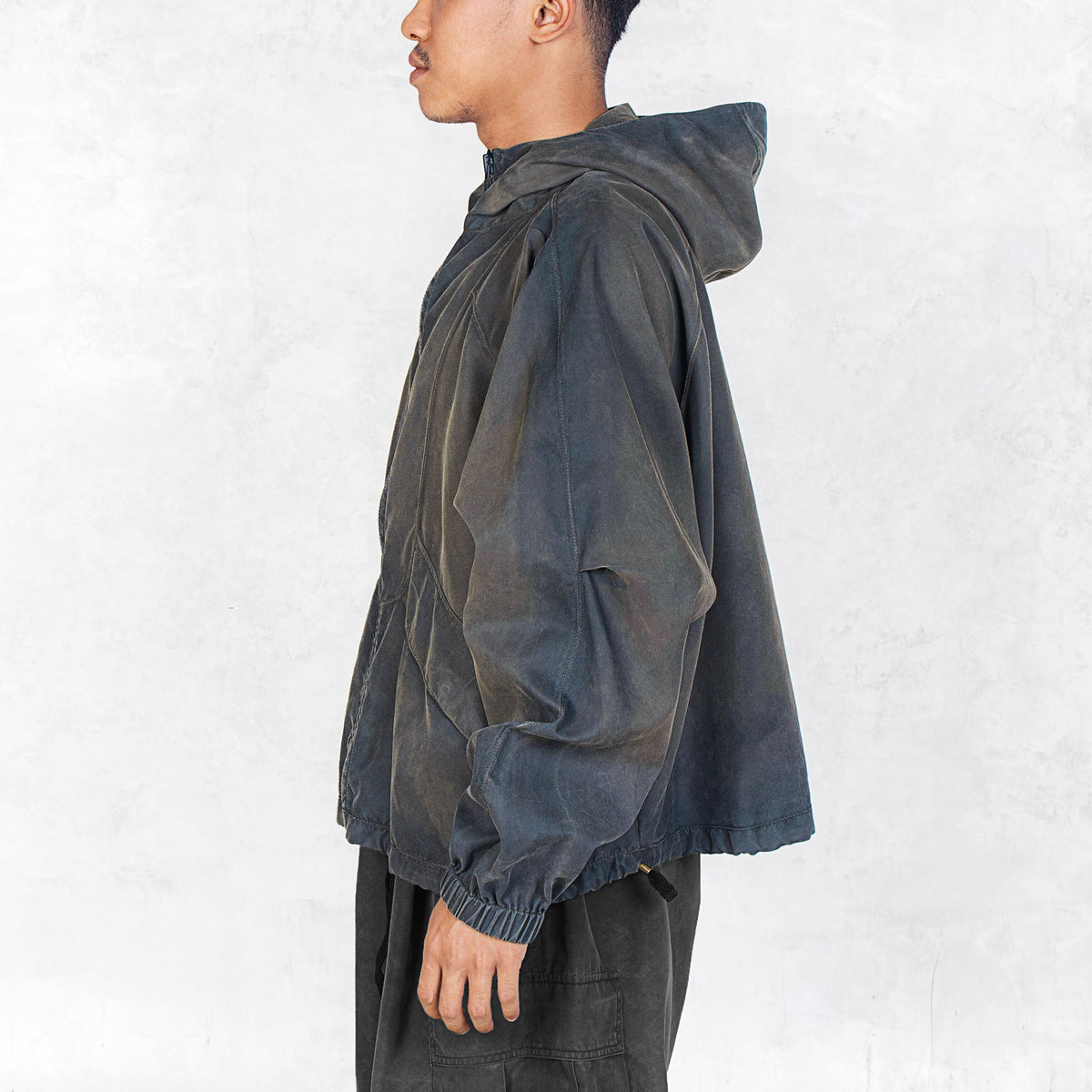 Person facing sideways wearing a dark olive zip up hooded jacket against a light grey background.