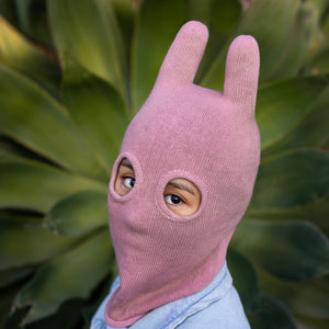 Person wearing a pink balaclava mask with ears standing in front of a plant with big green leaves 