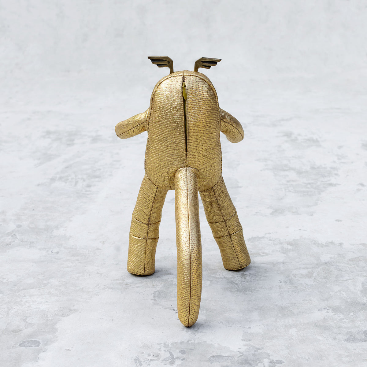 Gold leather and brass plush toy facing backwards on a textured white background
