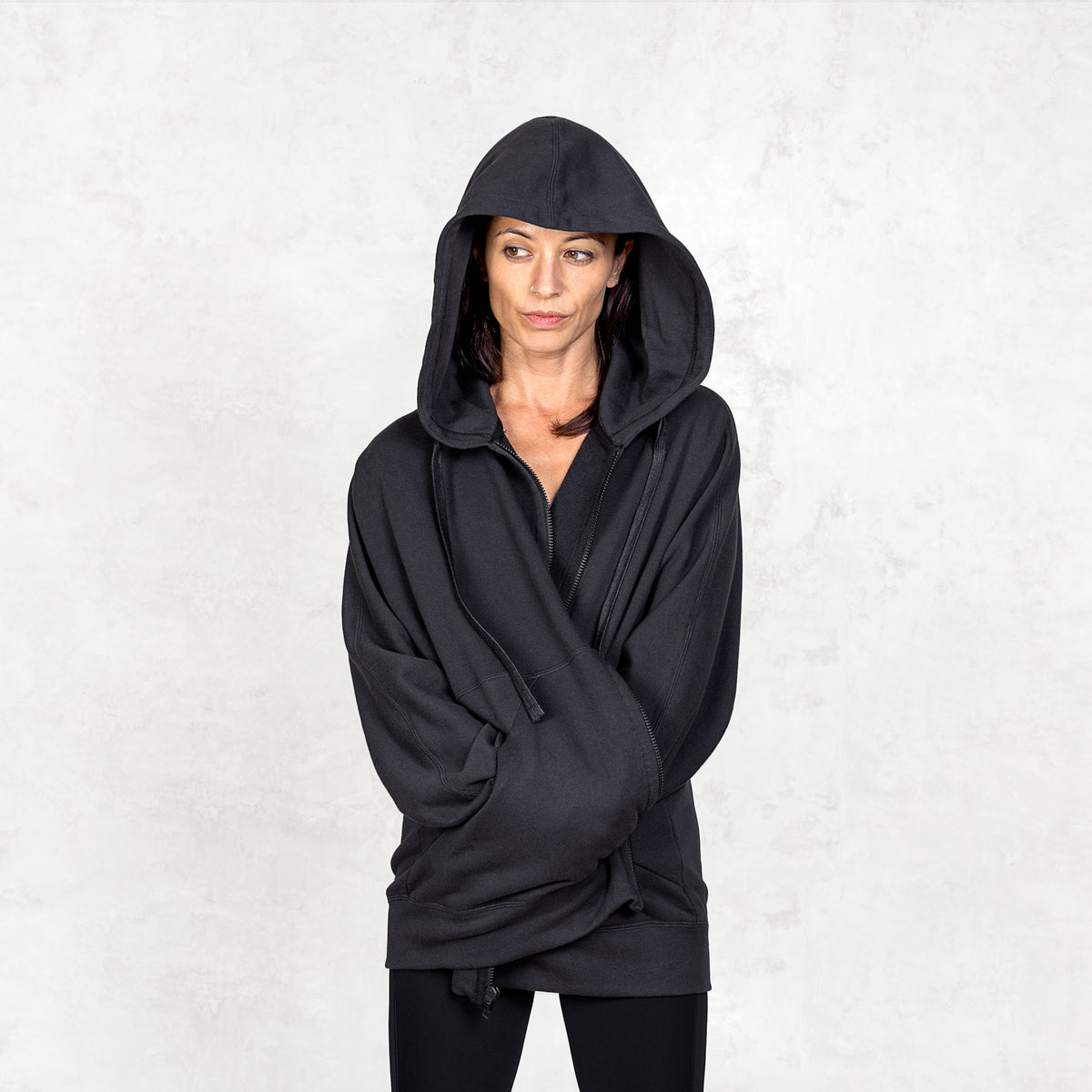 Person wearing a black hooded garment against a white background with their arms wrapped in front of them