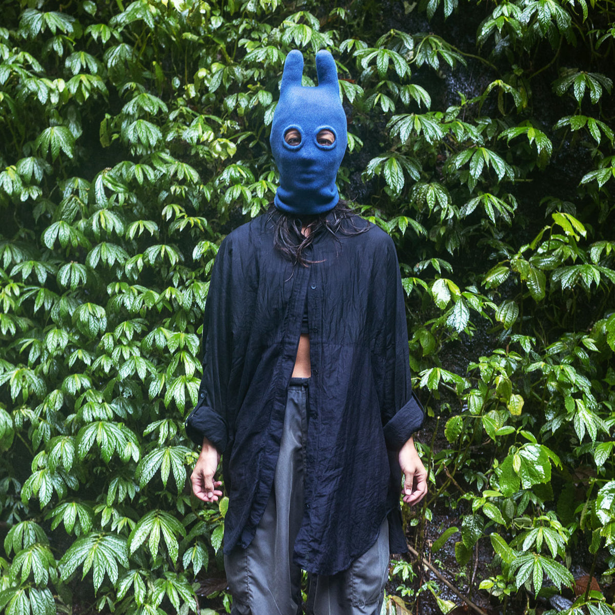 Person wearing a blue bunny mask standing in front of green foliage