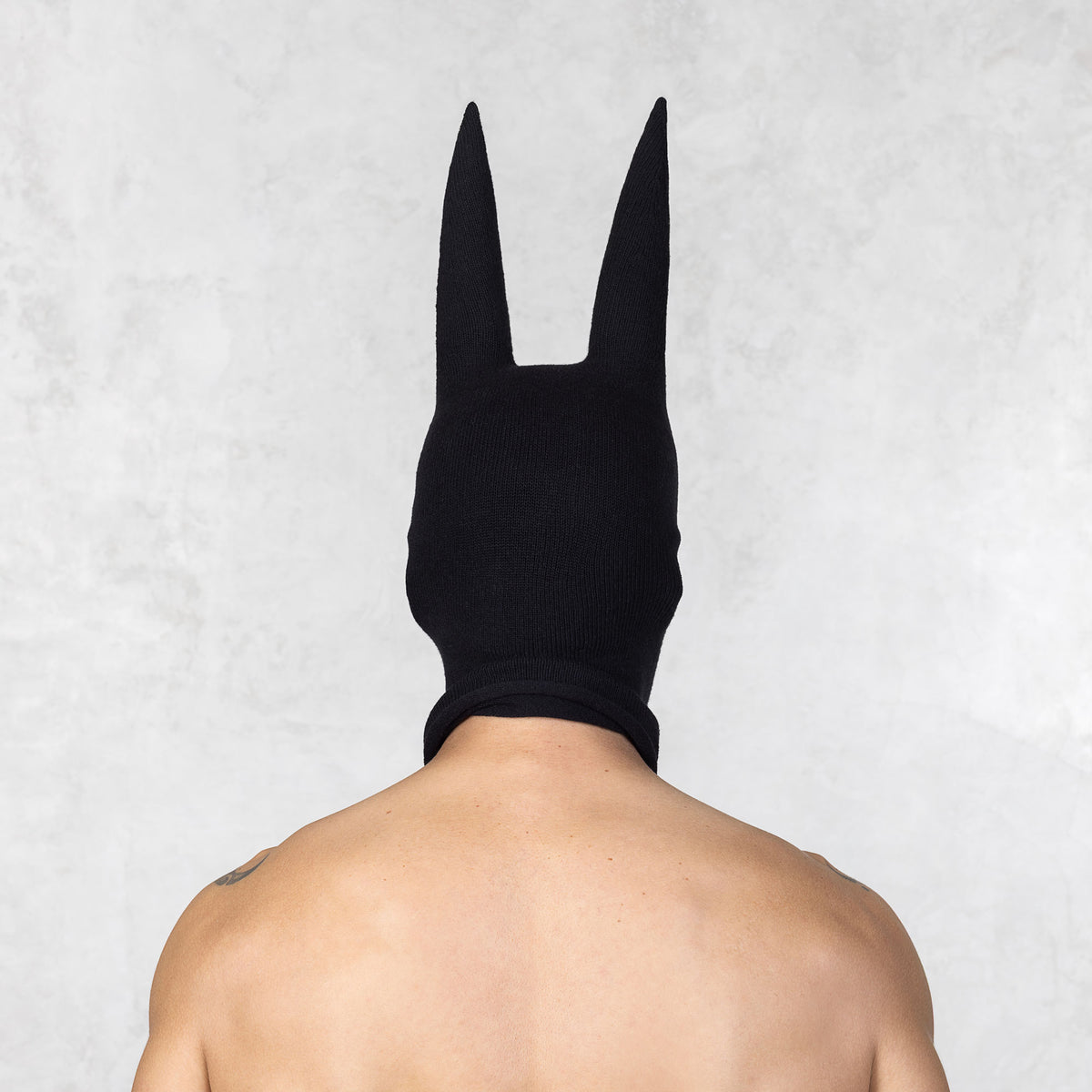 Person with tattoos wearing a black mask with ears facing backwards, standing against a plain background