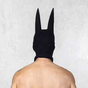 Person with tattoos wearing a black mask with ears facing backwards, standing against a plain background