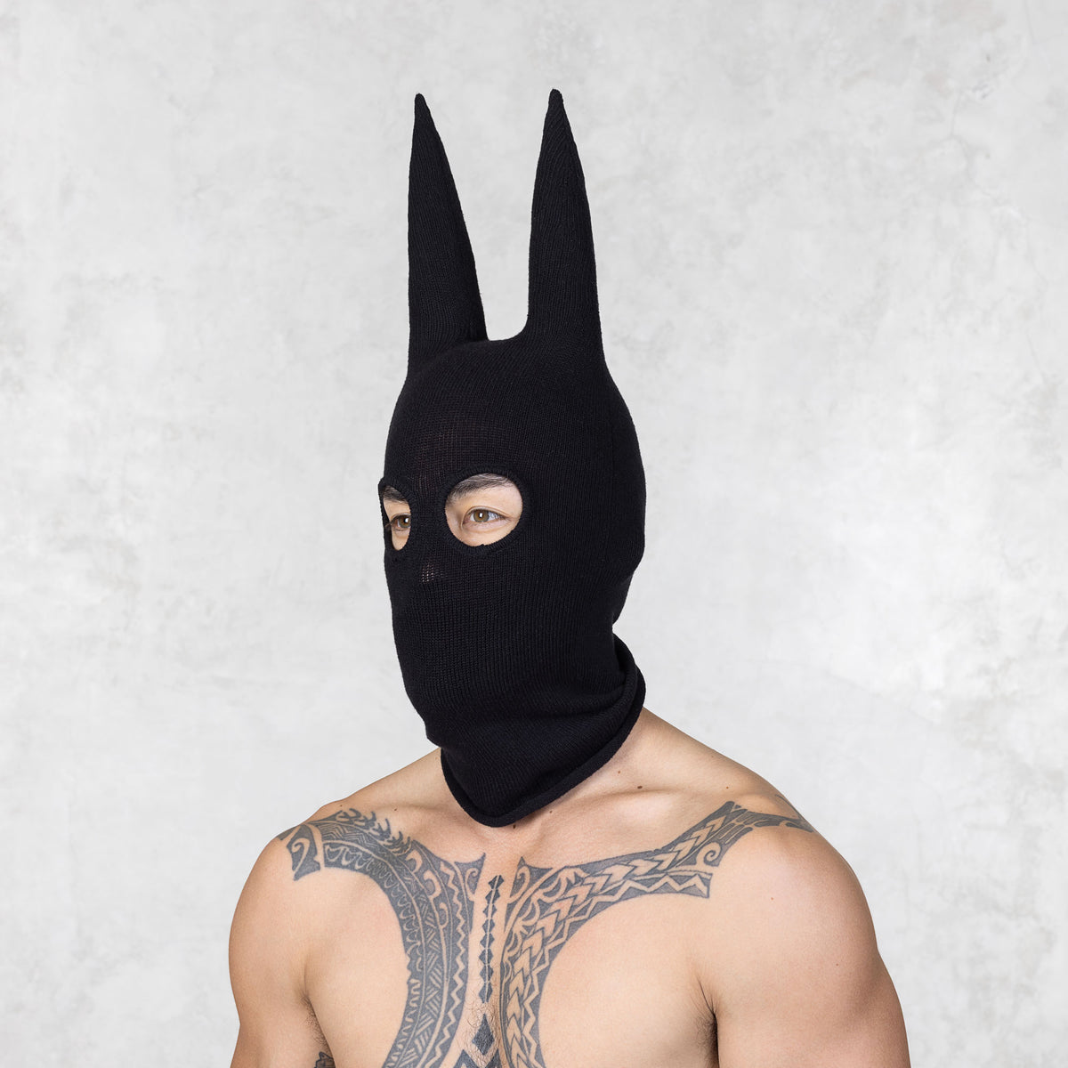 Person with tattoos wearing a black mask with ears looking away from the camera standing against a plain background