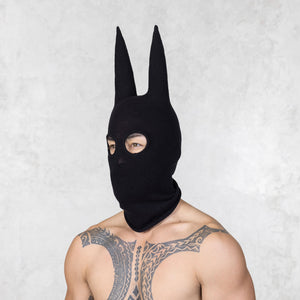 Person with tattoos wearing a black mask with ears looking away from the camera standing against a plain background