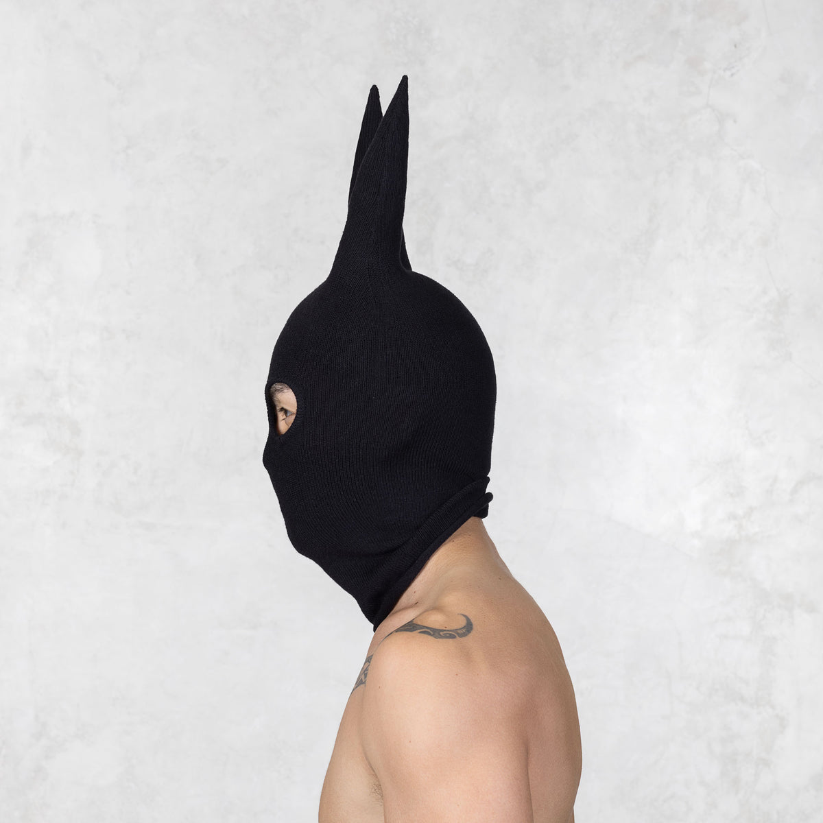 Person with tattoos wearing a black mask with ears facing sideways, standing against a plain background