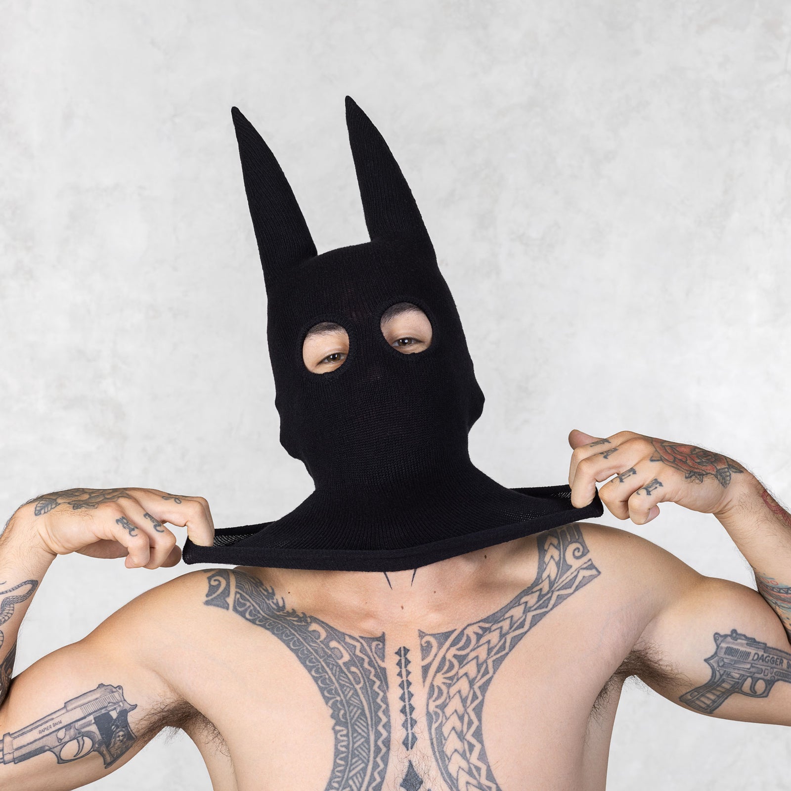Person with tattoos wearing a black mask with ears pulling the sides out, standing against a plain background