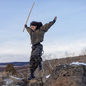 Person wearing dark baggy clothing holding a large wooden stick jumping off of a rock in a natural landscape