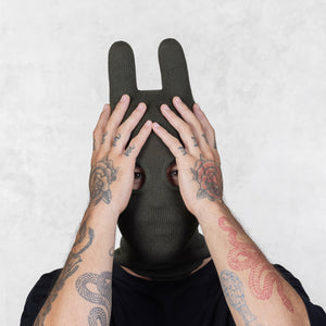 Person wearing a dark green knit balaclava with tattoos on arms and their hands over their face against a light background
