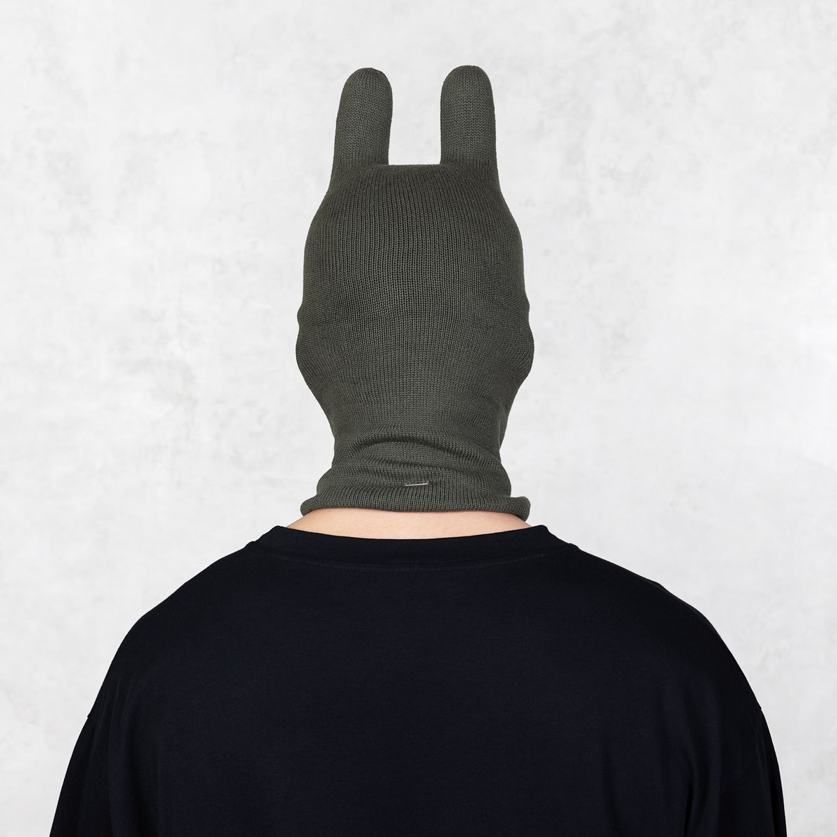 Person wearing a dark green balaclava facing backwards on a light gray background
