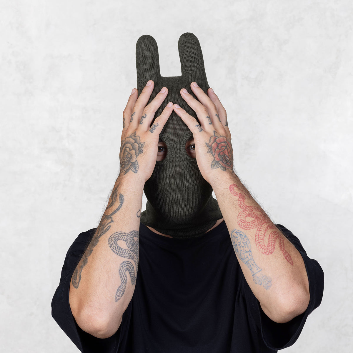 Person wearing a dark green balaclava covering their face with their tattooed arms on a light gray background