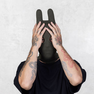 Person wearing a dark green balaclava covering their face with their tattooed arms on a light gray background