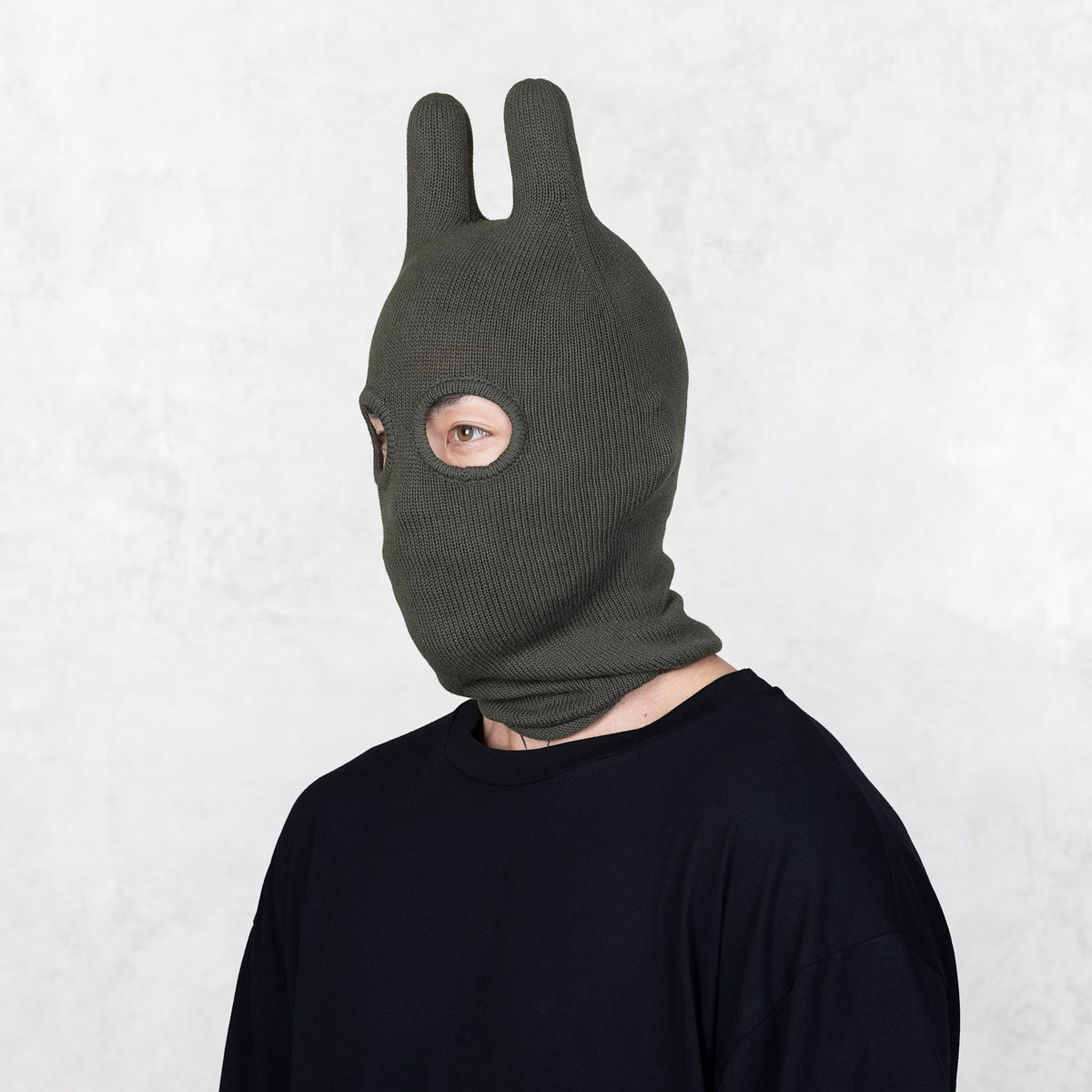 Person wearing a dark green balaclava facing off to the side on a light gray background