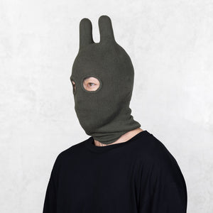 Person wearing a dark green balaclava facing off to the side on a light gray background