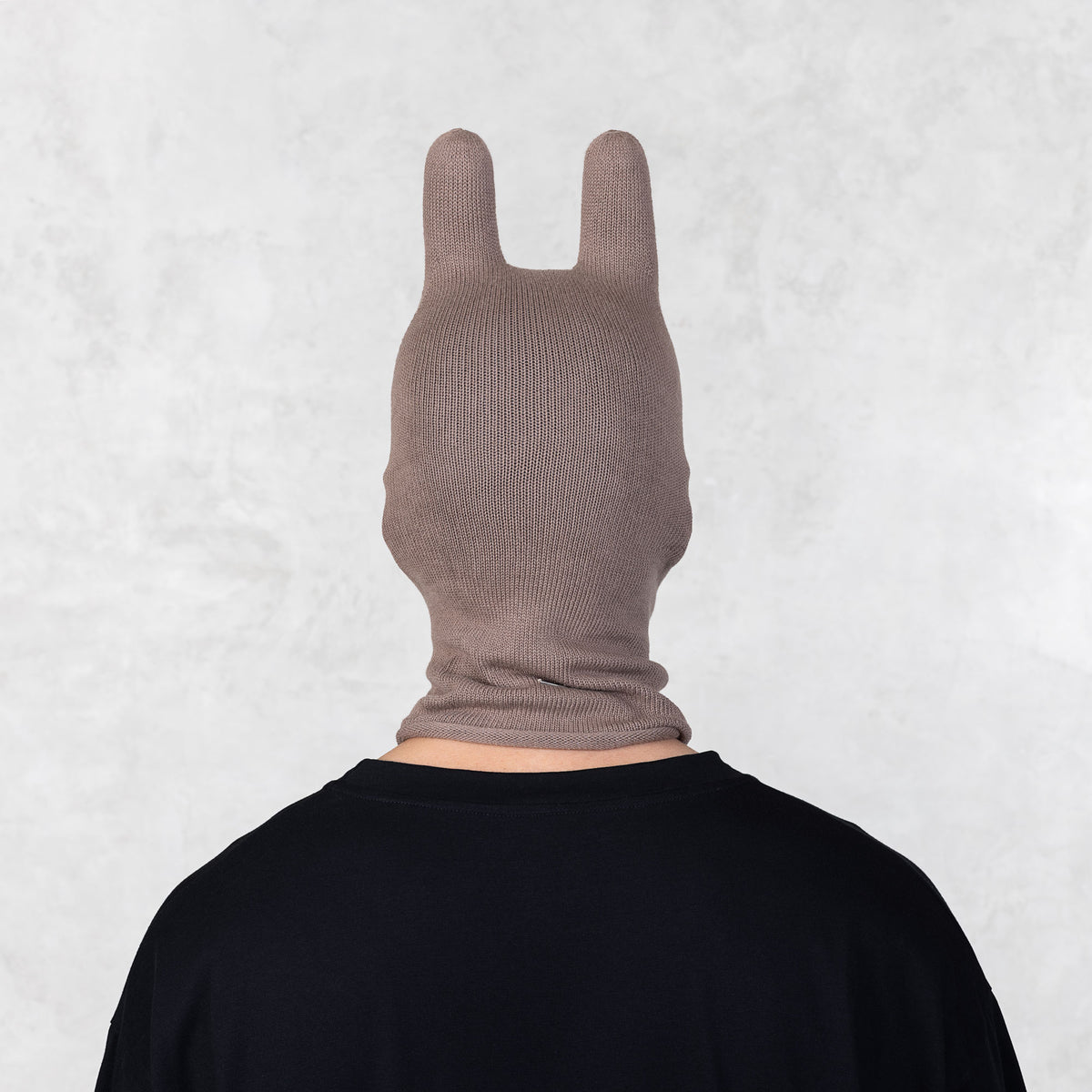 Person wearing a brown balaclava facing backwards on a light gray background