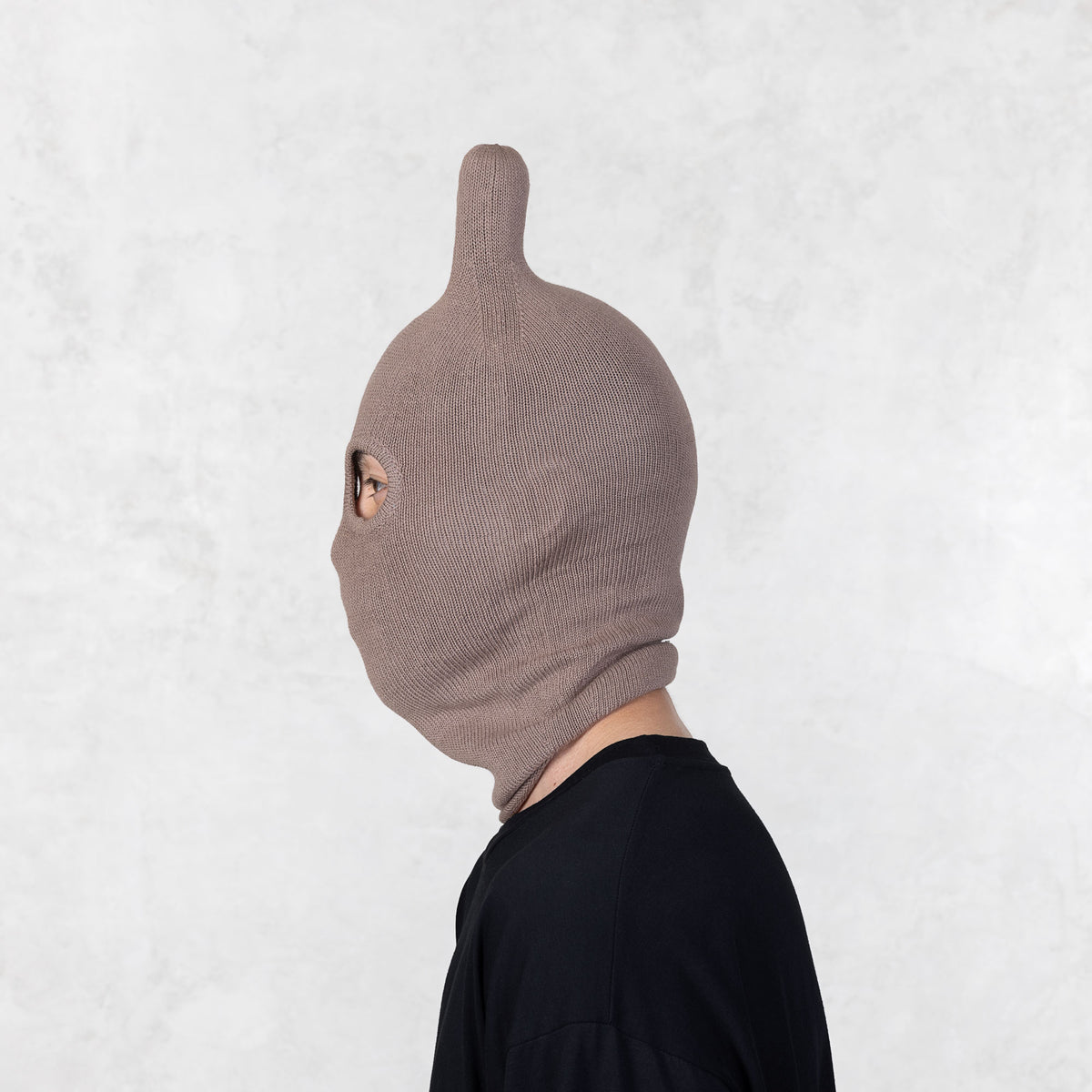 Person wearing a brown knit balaclava mask facing sideways against a white background