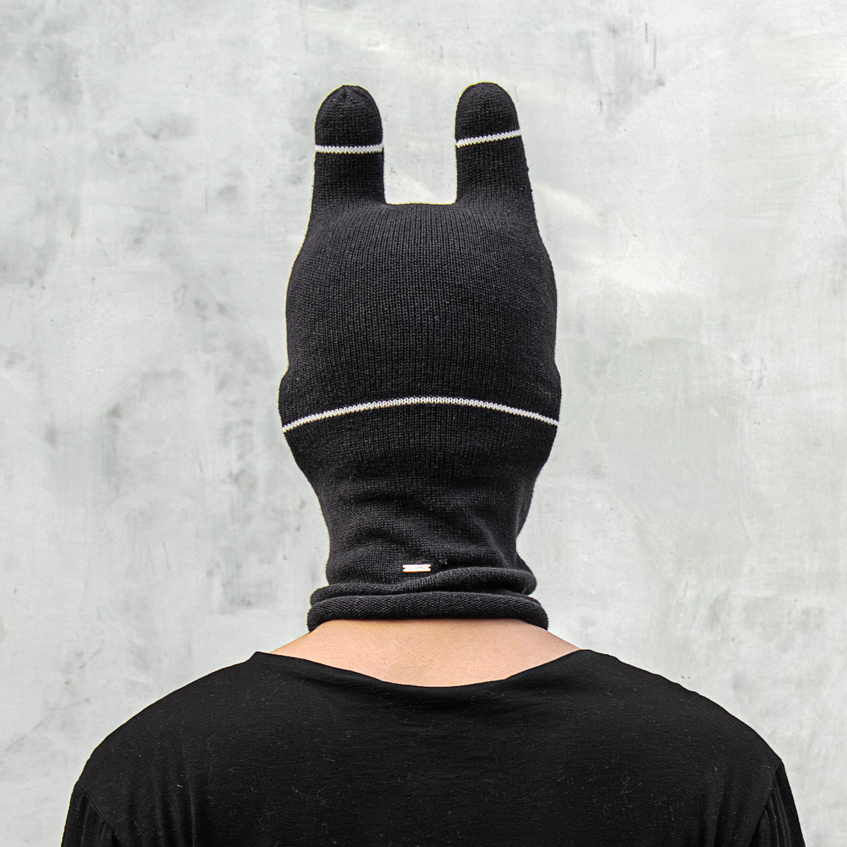 Person wearing a black balaclava with white stripes and ears against a gray background 