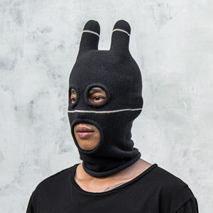 Person wearing a black balaclava mask with withe stripes and eye and mouth holes against a light gray background 