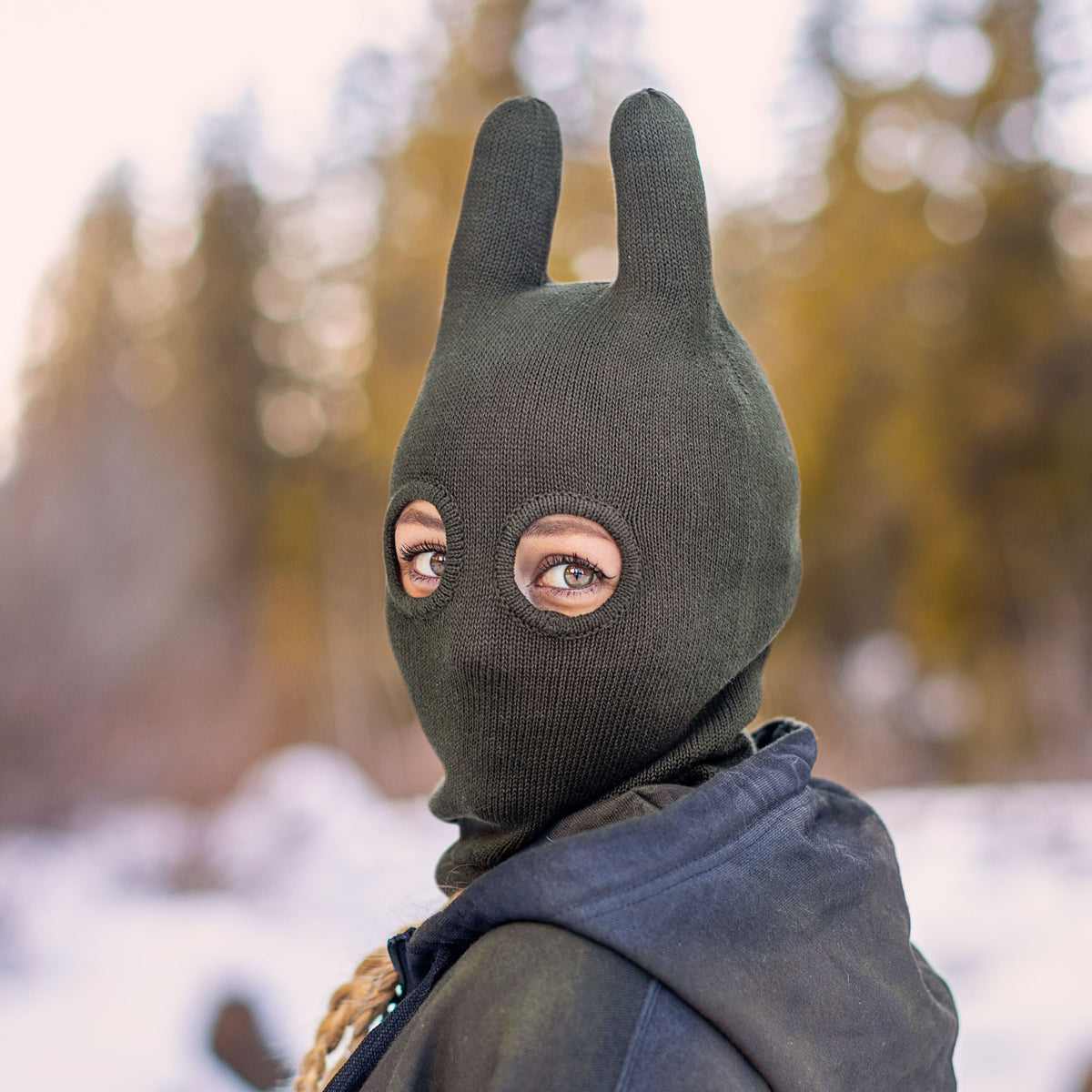Person wearing a dark olive knit balaclava mask with a blurred nature background