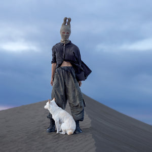 Person wearing an olive green balaclava with wavy horns, a black button up shirt unbuttoned on the bottom half, green baggy pants, and black boots standing on a sandy surface with a small white dog sitting next to them