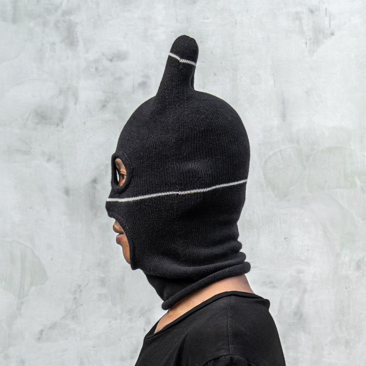 Person wearing a black balaclava mask with ears, eyes holes, and a mouth hole against a light gray background 