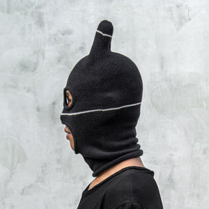 Person wearing a black balaclava mask with ears, eyes holes, and a mouth hole against a light gray background 
