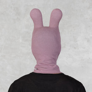 A person viewed from behind wearing a ribbed knit bunny-ear hood or balaclava in dusty pink over a black shirt, against a textured light gray background.