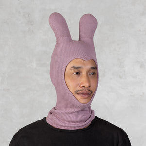 A person wearing a dusty pink ribbed knit balaclava with bunny ears, paired with a black shirt, set against a textured gray background.