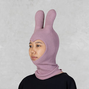 A person wearing a light pink ribbed knit balaclava-style hood with two upright bunny ears, shown against a light gray textured background.