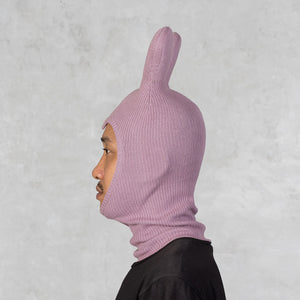 A person in profile wearing a light pink ribbed knit hood with a tall upright bunny ear, shown against a textured light gray background.