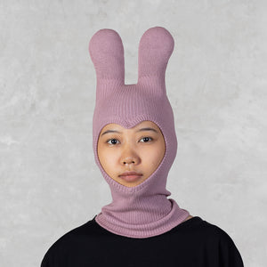 A person wearing a light pink ribbed knit balaclava hood with long ears, paired with a black top, against a textured light gray background.