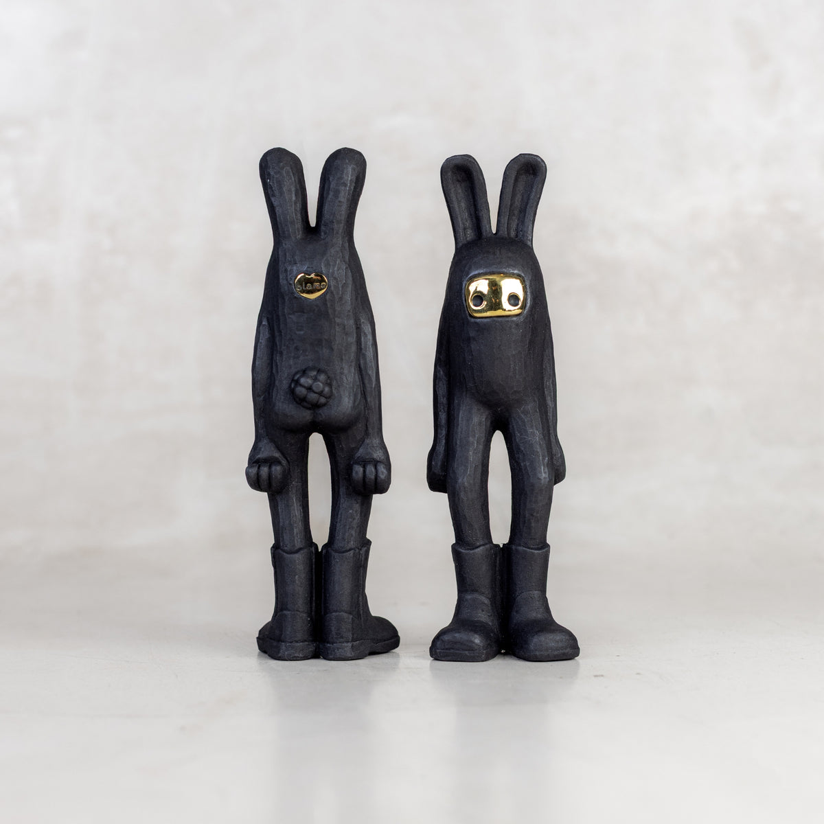 Two black rabbit-like figurines facing forward and backwards with a brass mask face on a white background