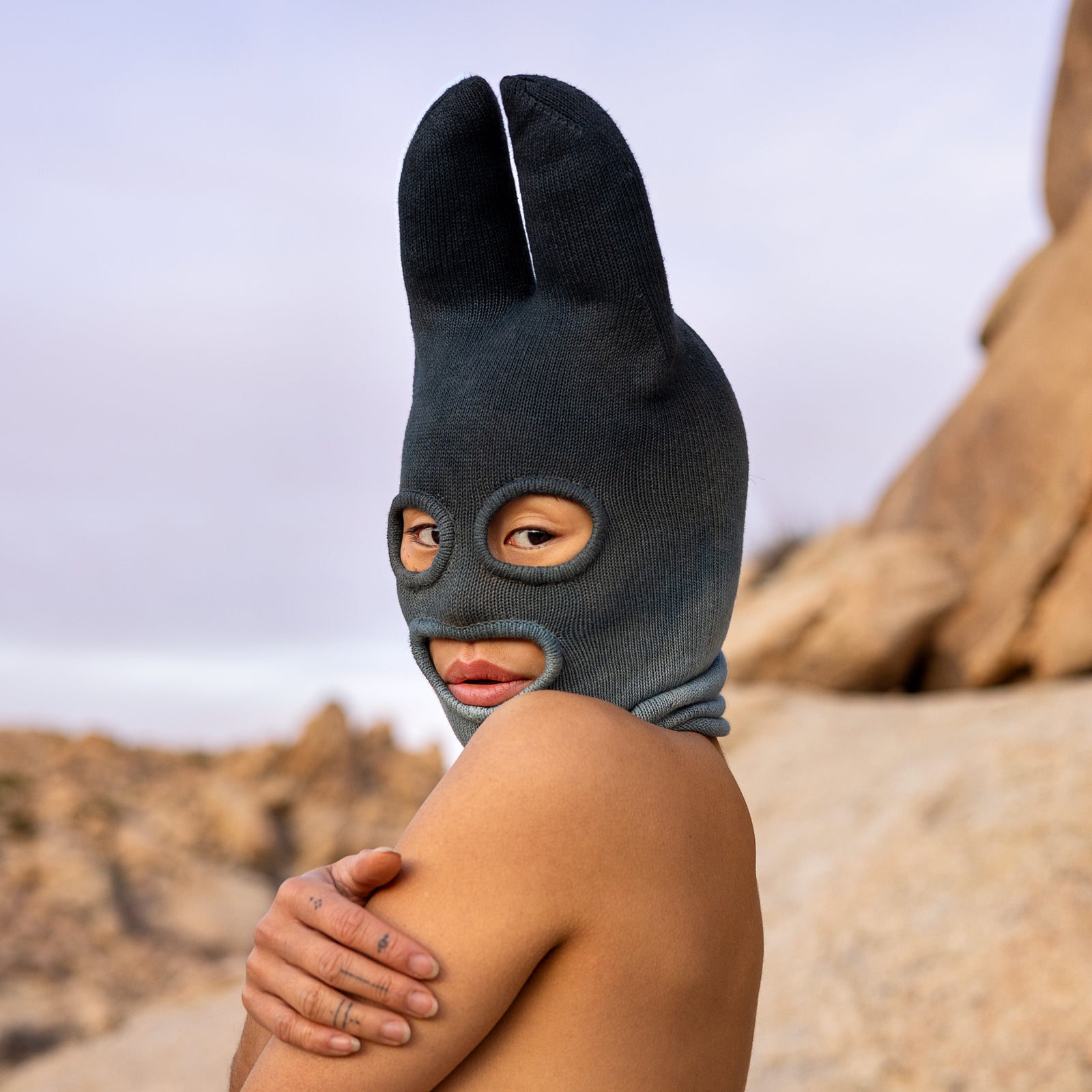 A person from the shoulders up wearing a cotton Knit Bat balaclava in faded indigo against a natural landscape